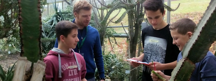 Student mit drei Schuelern im Xerophytenhaus. Schueler untersuchen Aloeblatt.