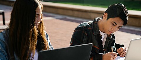 Zu sehen sind zwei junge Menschen, die draußen an Laptops arbeiten