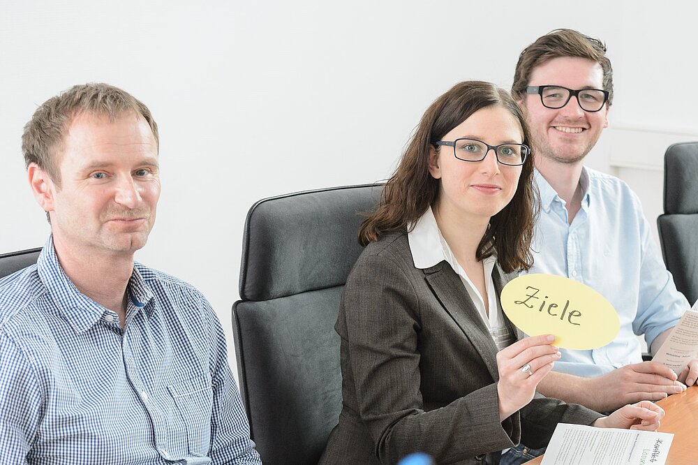 Two smiling men and one smiling woman with a card that says "goals"