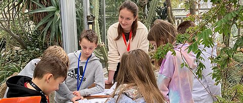 Das Foto zeigt eine Gruppe von Schüler:innen einer Grundschule mit einer Studentin bei der Stationenarbeit im Tropenschauhaus des Botanischen Gartens.