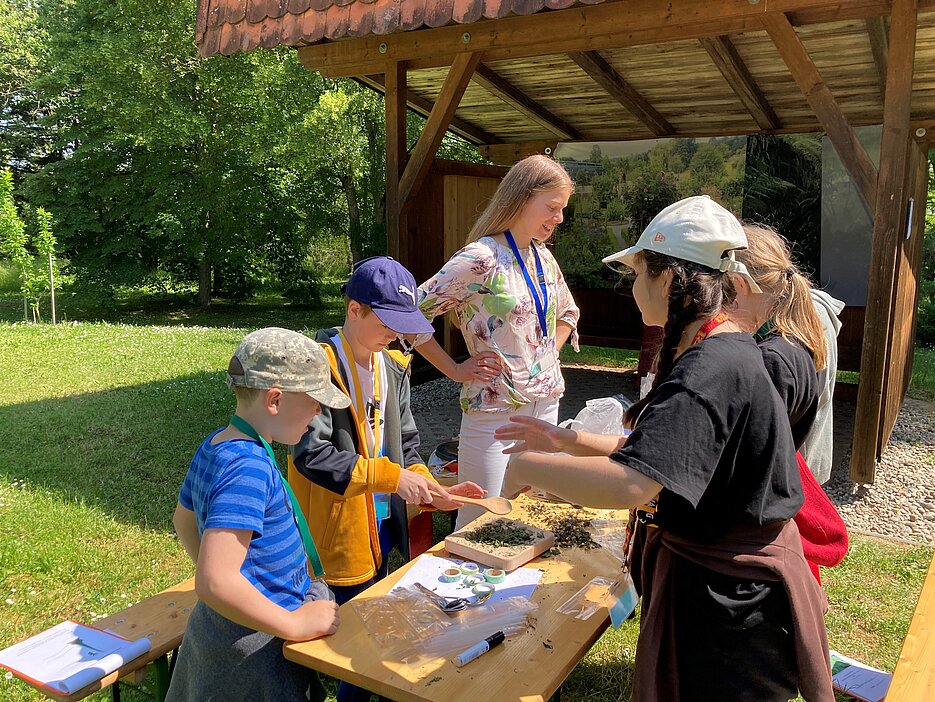 Das Foto zeigt eine Studentin mit einer Gruppe von Schüler:innen beim Herstellen von Brennnesselsalz an einer Station des Workshops im Freiland des Botanischen Gartens.