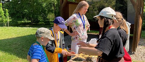 Das Foto zeigt eine Studentin mit einer Gruppe von Schüler:innen beim Herstellen von Brennnesselsalz an einer Station des Workshops im Freiland des Botanischen Gartens.