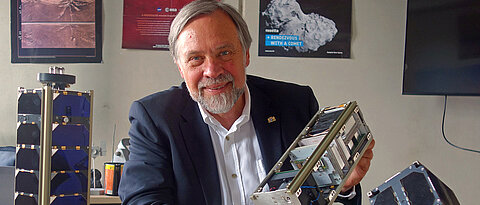 Klaus Schilling mit den Satelliten, die sein Berufsleben geprägt haben. UWE und NetSat im Vordergrund; auf den Postern im Hintergrund HUYGENS und ROSETTA, die er während seiner Tätigkeit in der Industrie mit realisieren half.