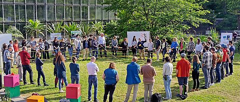 Das Foto zeigt auf einer Wiese einen Menschenkreis. Die Menschen halten ein gelbes Seil in den Händen. Das Seil spannt sich in Netzform über die Kreismitte.