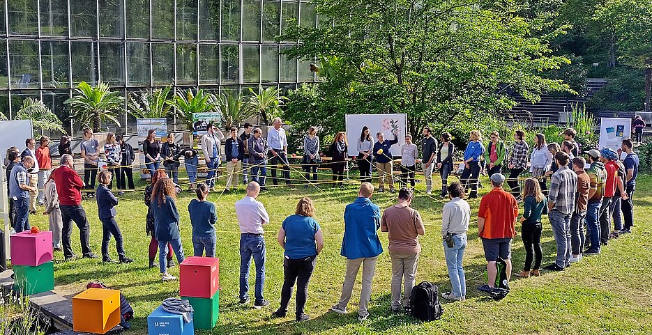 Das Foto zeigt auf einer Wiese einen Menschenkreis. Die Menschen halten ein gelbes Seil in den Händen. Das Seil spannt sich in Netzform über die Kreismitte.