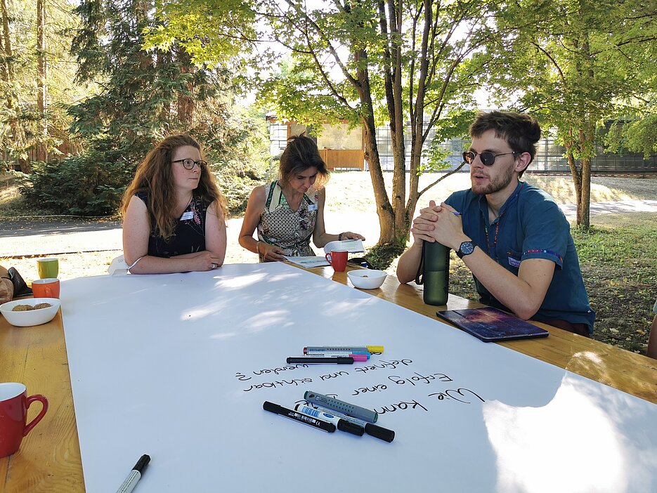 In einem World Café setzten sich die Teilnehmenden an drei Stationen mit Fragen zur Implementierung einer BNE in der Breite der Lehre auseinander. Das Foto zeigt eine der drei Stationen im Botanischen Garten, wo Dozierende an einem Tisch sitzend diskutieren und Ideen aufschreiben. An der Station im Foto ging es um die Definition und Sichtbarkeit einer erfolgreichen Nachhaltigkeitsbildung an der Uni. (Foto: Anna Dävel)