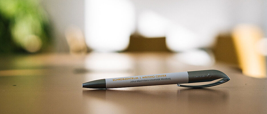 Symbolbild Schreibberatung (Ein Stift mit dem Logo des Schreibzentrums der Universität Würzburg liegt auf einem Tisch.)