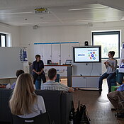 Personen sitzen zusammen und sprechen. Ein Bildschirm mit dem Ideenlabor-Logo steht im Raum.
