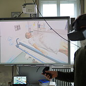 Ein Mann mit VR-Brille hält zwei Controller in der Hand. Hinter ihm ein Bildschirm mit einer Simulation von einem Behandlungszimmer mit Patienten.