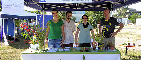 2 Studierende, Koordinatorin Dr. Kerstin Bissinger und Kustos Dr. Gerd Vogg am Stand des Botanischen Gartens, auf dem Tisch steht ein Blumenstraus und verschiedene fleischfressende Pflanzen