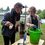 Zwei Mädchen arbeiten mit einem Filterversuch. In einer Glassäule befinden sich verschiedene Bodenschichten. Die Kinder geben verschmutztes Wasser auf die oberste Schicht und betrachten das austretende saubere Wasser.