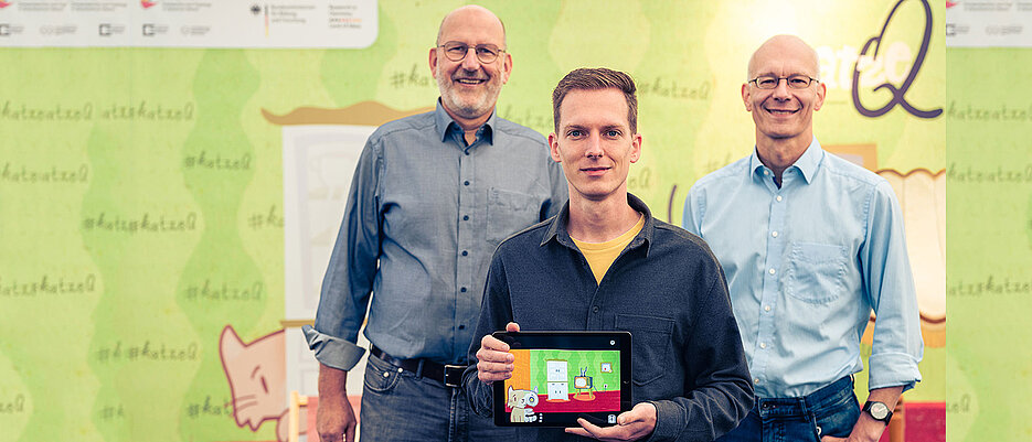 The two spokespersons of the Cluster of Excellence ct.qmat, Prof. Ralph Claessen (l.) and Prof. Matthias Vojta (r.), together with app designer Philipp Stollenmayer (center), present their joint project, the game app "Kitty Q". 