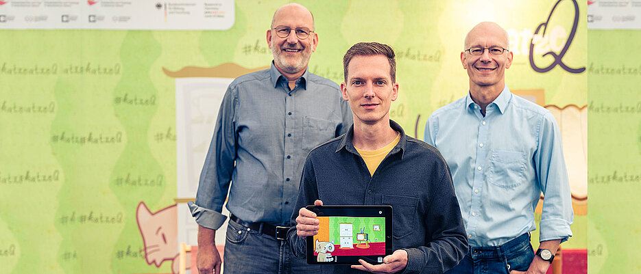 Die beiden Sprecher des Exzellenzclusters ct.qmat, Ralph Claessen (l.) und Matthias Vojta (r.), präsentieren mit dem App-Designer Philipp Stollenmayer ihr gemeinsames Projekt, die Spiele-App „Katze Q“.