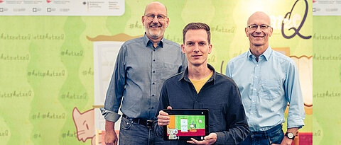 The two spokespersons of the Cluster of Excellence ct.qmat, Prof. Ralph Claessen (l.) and Prof. Matthias Vojta (r.), together with app designer Philipp Stollenmayer (center), present their joint project, the game app "Kitty Q". 