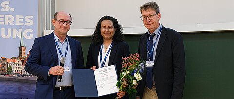Die diesjährige DGHM-Hauptpreisträgerin Cynthia Sharma bei der Preisverleihung mit DGHM-Präsident Prof. Jan Buer (r) und Prof. Klaus Pfeffer, dem Vorsitzenden der DGHM-Stiftung.