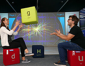 In der interaktiven Wissenschaftsausstellung Touch Science sitzen zwei Besucher auf Kissen, die zwei verschiedene Elementarteilchen symbolisieren. Im Hintergrund ist das Ereignisbild einer Teilchenkollision im ATLAS-Detektor am CERN zu sehen. Mit solchen 