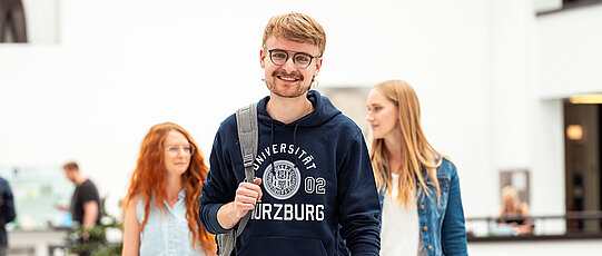 Person schreibt mit Stift, Das Logo der Universität ist verschwommen im Hintegrund zu sehen.