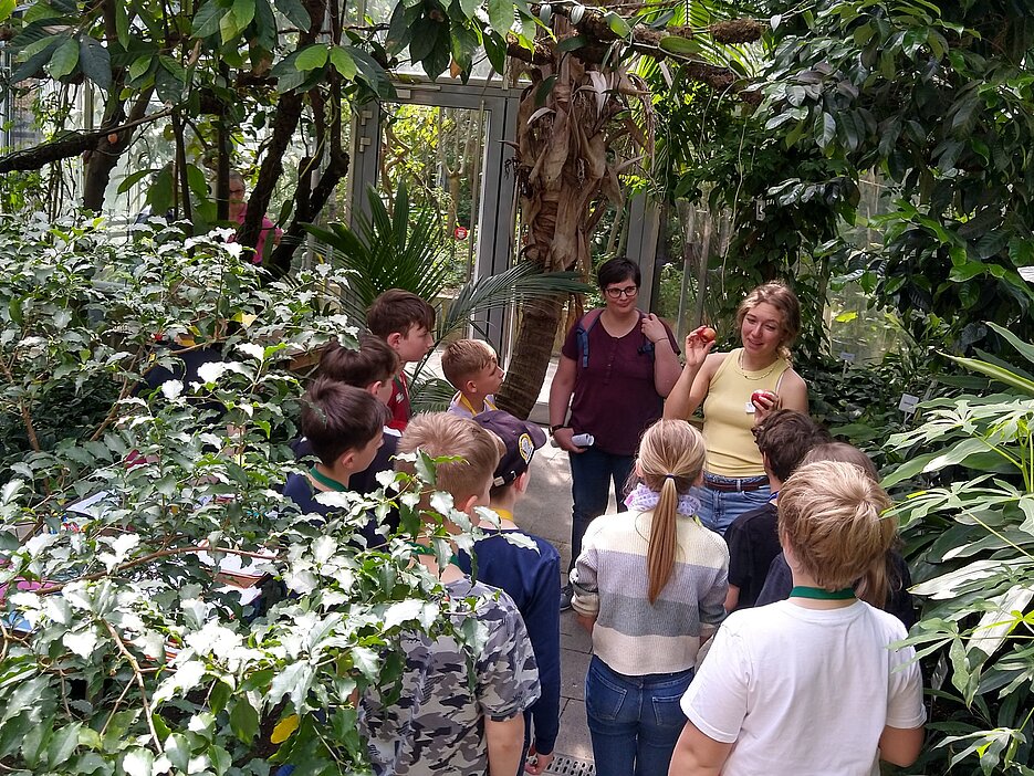 Das Foto zeigt eine Studentin mit einer Gruppe von Schüler:innen an der Workshop-Station zu saisonalen und regionalen Früchten im tropischen Nutzpflanzenhaus des Botanischen Gartens.