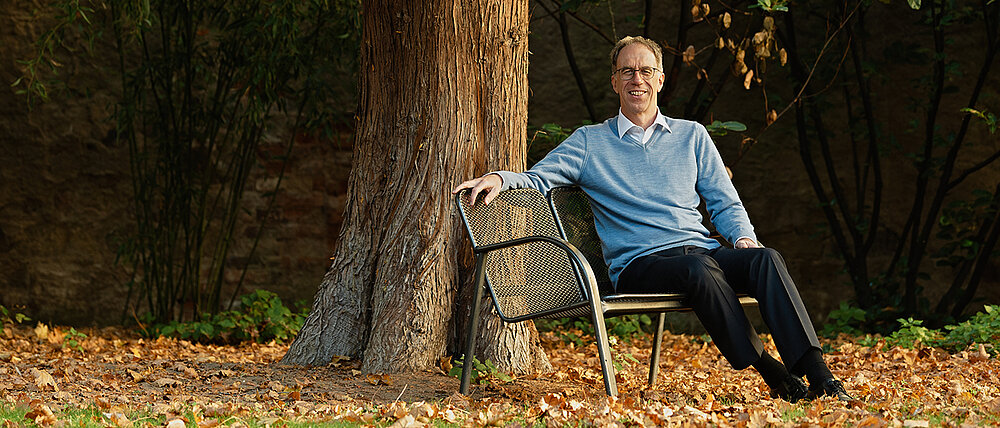 Präsident Pauli sitzt auf einer Bank unter einem Baum umgeben von herbstlichem Laub.