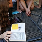 Kinder sitzen um ein Tablet und ein aufgeschlagenes Buch herum. Im Buch steht ein Text auf Hebräisch. Auf der anderen Buchseite steht ein einziges hebräisches Wort und daneben ein Post-it mit der Aufschrift "Wichtig!" 