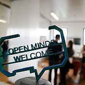 Im Vordergrund ist die Figur mit dem Text "Open Minds Welcome" und im Hintergrund die Teilnehmer zu sehen.