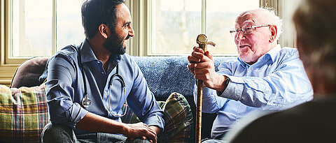 Young male doctor and senior man sitting on sofa and smiling during home visit