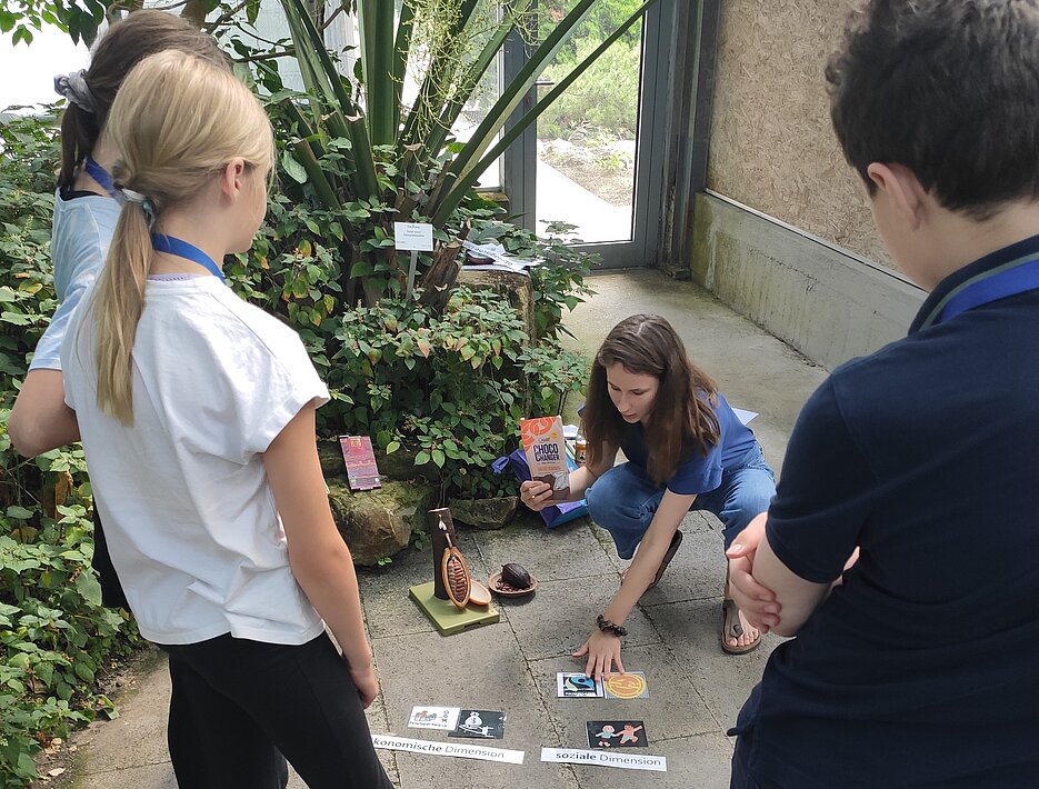 Die Schüler:innen stehen im Kreis und die Studentin legt verschiedene Begriffskarten zum Thema der Nachhaltigkeit in der Mitte aus.