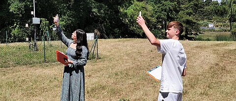 An der dritten Station des Workshops setzten sich die Schüler:innen mit dem Zusammenhang zwischen Wetter und Klima auseinander. Dazu nahmen sie vor Ort auch eigene Messungen vor. Das Foto zeigt eine Schülerin und einen Schüler, die mithilfe von Messgeräten die Temperatur am Standort der Station 3 im Botanischen Garten ermitteln. Im Hintergrund ist die Wetterstation im Botanischen Garten zu sehen. (Foto: Anna Dävel)