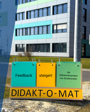 Ein Aufsteller mit der Aufschrift "Feedback steigert die Selbstwirksamkeit von Studierenden" steht vor einem Uni-Gebäude.