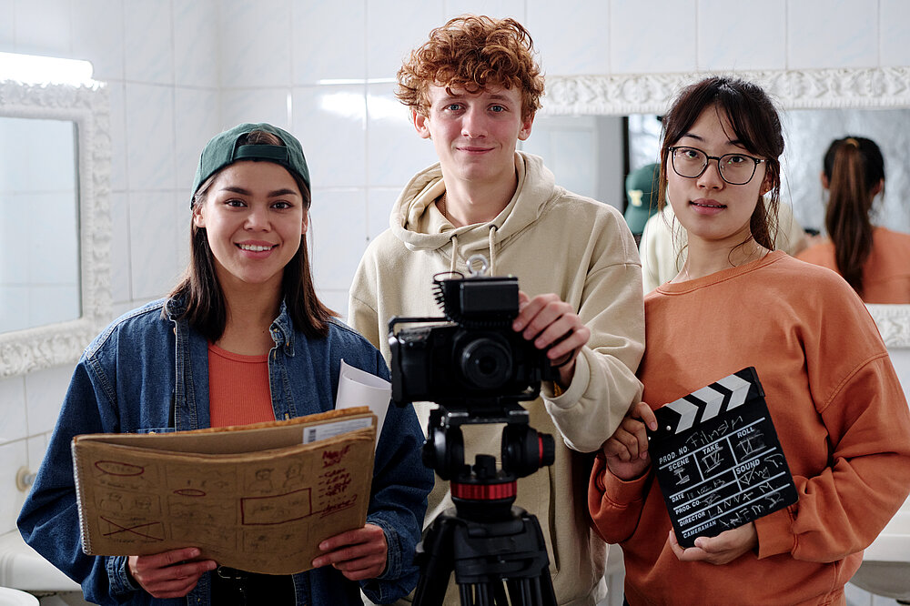 Portrait of three young people holding film equipment and smiling warmly at camera in studio setting while standing near large mirror in background