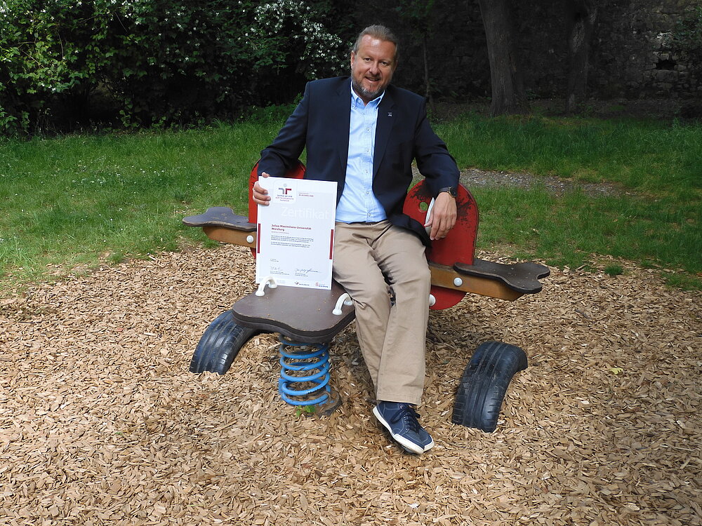 Baumann hält die Urkunde des Zertifkats Familiengerechte Hochschule in der Hand und sitzt auf einem Spielplatz.