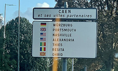 Schild an einer Straße, darauf aufgelistet sind alle Partnerstädte von Caen. Würzburg steht an erster Stelle.