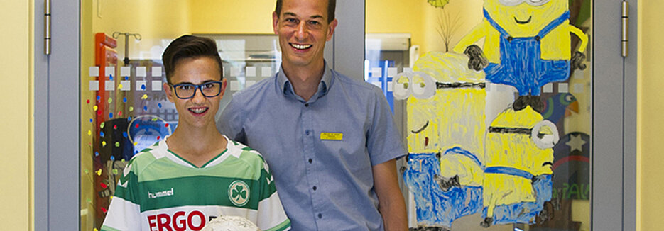 Der ehemalige MDS-Patient Lucas Melchner und Privatdozent Dr. Matthias Wölfl vor der Stammzelltherapie-Station „Schatzinsel“ der Würzburg Universitäts-Kinderklinik. (Foto: Arnika Hansen/Uniklinikum Würzburg)