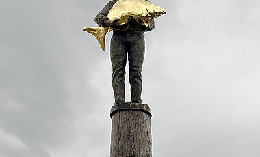 Das Kunstwerk „Der Diez“ in Dießen am Ammersee zeigt einen Mann, der einen goldenen Karpfen hält. Das Denkmal symbolisiert die Bedeutung der Fischerei für die Menschen der Region.