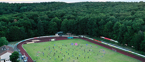 So sieht es aus, wenn die Das Bild zeigt die Teilnehmer des Global Village unter Pandemiebedingungen feiern: 100 Studierende in einem Kreis mit einem Durchmesser von 70 Metern, jeweils zwei Meter voneinander entfernt auf dem  Sportplatz der TG Höchberg.
