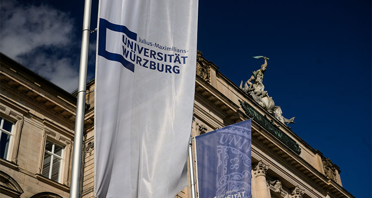 Das Hauptgebäuder der Uni Würzburg am Sanderring. Vor dem Gebäude sind Fahnen mit dem Logo der Uni Würzburg gehisst.